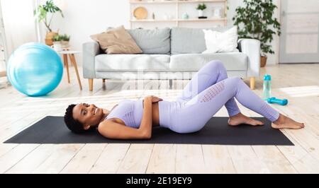 Giovane afroamericana sdraiata sul tappeto yoga in soggiorno, riposandosi dopo un intenso allenamento, panorama Foto Stock