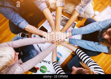 I giovani dipendenti motivati sono seduti sul pavimento dell'ufficio in cerchio e tengono insieme le mani in mezzo. Lavoro di squadra e concetto di collaborazione. Foto Stock