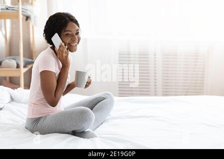 Allegra donna nera seduta a letto, che parla sul telefono cellulare Foto Stock