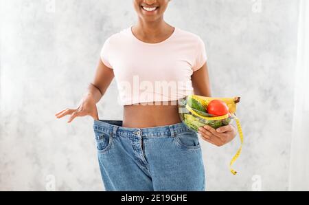 Vista ritagliata della donna nera con ciotola di frutta, verdure e nastro di misurazione che indossa jeans oversize su sfondo chiaro Foto Stock