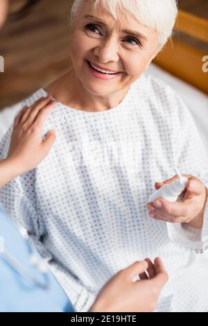 donna anziana sorridente che tiene lo spray nasale vicino all'infermiere che tocca sentire spalla su primo piano sfocato Foto Stock