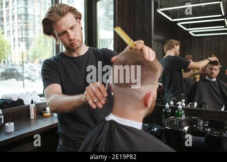 Giovane barbiere professionista che guarda messo a fuoco, dando un taglio di capelli ad un cliente Foto Stock