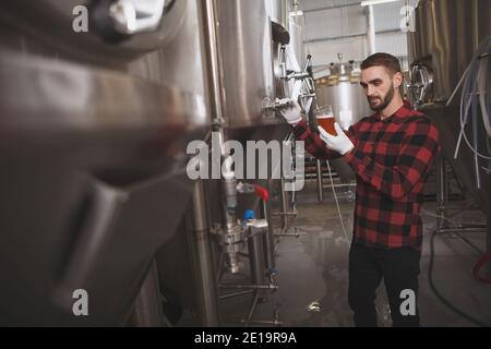 Birreria maschile che assaggia birra appena prodotta presso la sua fabbrica di birra, vasche di metallo sullo sfondo, spazio copia Foto Stock