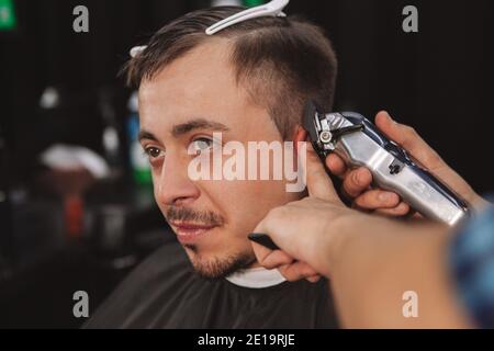 Primo piano di un uomo maturo che si diverti a ottenere un nuovo taglio di capelli presso il barbiere locale. Ritaglio di un barbiere professionale utilizzando il regolacapelli elettrico, dando Foto Stock