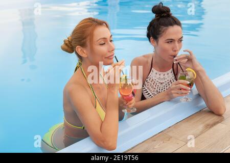 Due belle giovani donne che si gustano un cocktail a bordo piscina del loro hotel. Attraenti amici femminili che celebrano la vacanza estiva, bevendo cocktail i Foto Stock