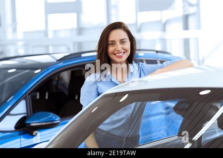 Macchina perfetta per me. Splendida giovane donna sorridente che si appoggia felicemente su un'auto presso la concessionaria auto locale Foto Stock