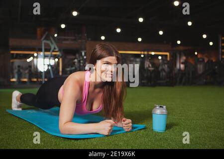 Bella donna in sovrappeso sorridente, facendo esercizio in planking in palestra, spazio di copia. Affascinante sportivo di dimensioni più che si allenano nello studio sportivo Foto Stock