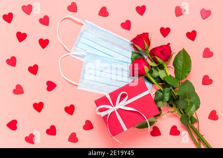 Rose rosse, confezione regalo e maschere facciali su sfondo rosa con molti cuori di legno. Festa delle madri, delle donne o dei San Valentino durante il coronavirus Foto Stock