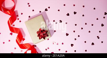 Confezione regalo e nastro rosso satinato sul lato su sfondo rosa. I piccoli cuori rossi sono sparsi. Spazio libero per il testo. Concetto regalo, vacanze, amore, va Foto Stock