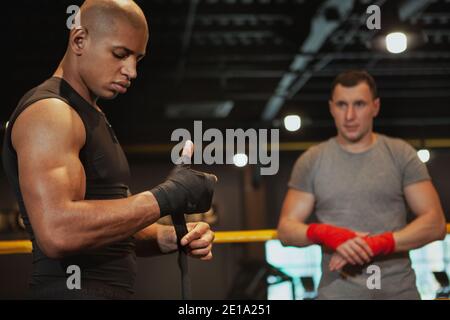 Bell'uomo africano muscoloso che si prepara per l'allenamento di boxe con il suo amico, copia spazio. Combattente di pugilato maschile che avvolge i pugni in bendaggi prima della boxe Foto Stock