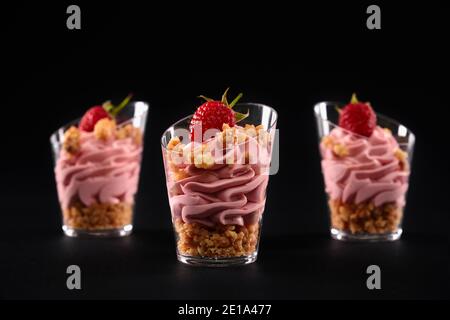 Vista panoramica del gustoso parfait con granola, decorato con lamponi sulla parte superiore e panna montata rosa. Dessert dolce fatto in casa a strati in tre piccoli bicchieri di fila isolati su sfondo nero. Foto Stock