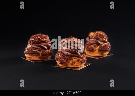 Tre deliziosi profiteroles croccanti freschi con ripieno cremoso di cioccolato marrone dolce. Closeup di gustosi eclair fatti in casa al forno isolato su sfondo nero. Concetto di dessert, cibo da ristorante. Foto Stock