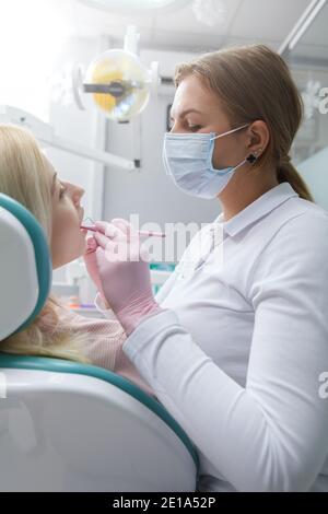 Scatto verticale di dentista femminile in maschera medica viso lavoro, esaminando i denti della paziente femminile Foto Stock
