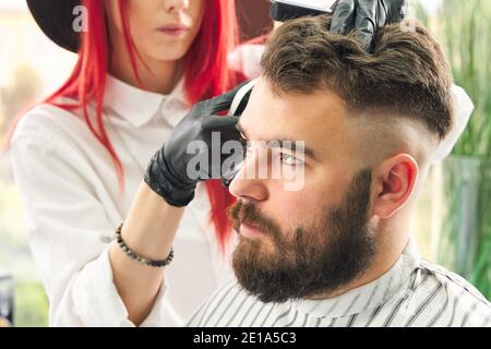 Il parrucchiere professionale utilizza un regolacapelli per frangere la barba un bell'uomo in barbiere Foto Stock