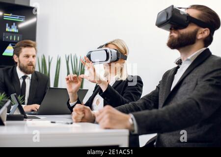 Realtà virtuale, tecnologia e concetto aziendale. Un team di tre designer o architetti professionisti di sesso maschile e femminile, che indossano cuffie per la realtà virtuale, che lavorano con nuove soluzioni virtuali Foto Stock