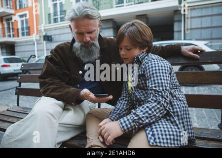 Uomo anziano che usa lo smartphone con l'aiuto del suo giovane nipote, seduto su una panchina in città Foto Stock