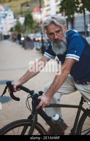 Immagine verticale di un uomo elegante senior con capelli grigi e la barba che cavalcano in bicicletta ghiaiata Foto Stock