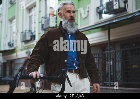 Bell'uomo elegante con bearded e capelli grigi che cammina per le strade della città con la sua bicicletta Foto Stock