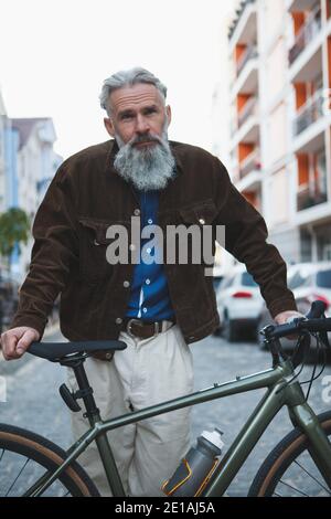 Ritratto verticale di un uomo con bearded alto e bello che indossa stile streetwear in piedi sulla strada della città con la sua bicicletta Foto Stock