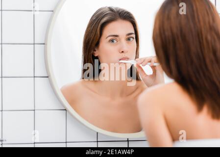 giovane donna che spazzolava i denti vicino allo specchio in bagno, in primo piano sfocato Foto Stock