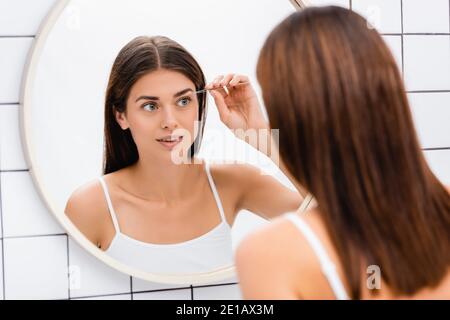 giovane donna in bianco singlet tweezing sopracciglia vicino specchio dentro bagno Foto Stock