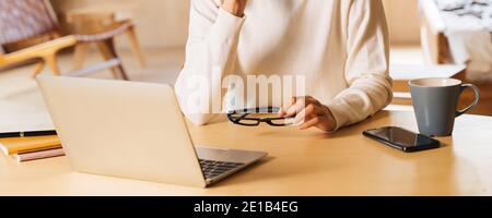 Giovane donna d'affari asiatica in felpa invernale con occhiali che usano il computer portatile sorridente. Donna freelance seduto al tavolo e che lavora da casa. Foto Stock