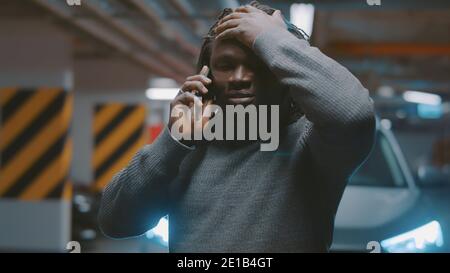 Uomo nero afro-americano sconvolto che parla sullo smartphone di fronte all'auto in garage. Foto di alta qualità Foto Stock