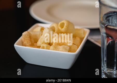 I Fryums serviti in una piccola ciotola bianca su un tavolo nero. Un bicchiere d'acqua in primo piano e un piatto bianco sullo sfondo. Foto Stock