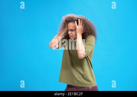 Esperienza di emicrania o mal di testa giovane donna afroamericana in piedi tenendo testa con le mani in t-shirt oliva isolato su sfondo blu. Umano Foto Stock