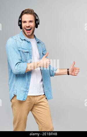 Felice giovane bell'uomo in camicia jeans che punta via in piedi su sfondo grigio con cuffie di grandi dimensioni Foto Stock