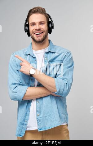 Felice giovane bell'uomo in camicia jeans che punta via in piedi su sfondo grigio con cuffie di grandi dimensioni Foto Stock