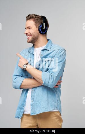 Felice giovane bell'uomo in camicia jeans che punta via in piedi su sfondo grigio con cuffie di grandi dimensioni Foto Stock