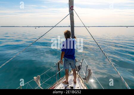 Le Grau-du-Roi (sud della Francia): Uomo in piedi sulla prua di una barca a vela che guarda la costa di fronte a Port-Camargue, uno dei più grandi porti turistici in Europa Foto Stock