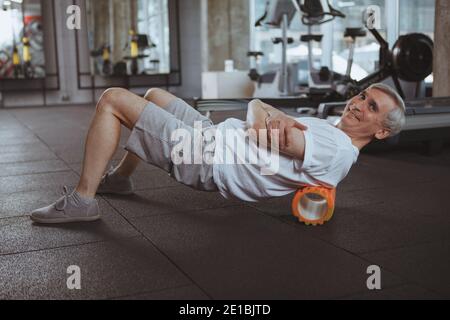 Uomo anziano allegro che sorride alla fotocamera, utilizzando il rullo in schiuma sulla schiena. Anziano sportivo che rilassa i muscoli della schiena dopo essersi allenato in palestra. Ma Foto Stock