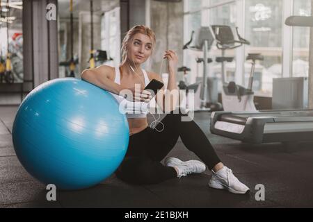 Splendida felice giovane sportiva sana utilizzando il suo smartphone in palestra, copia spazio. Bella donna atletica con capelli biondi che si rilassa dopo aver allenato w Foto Stock