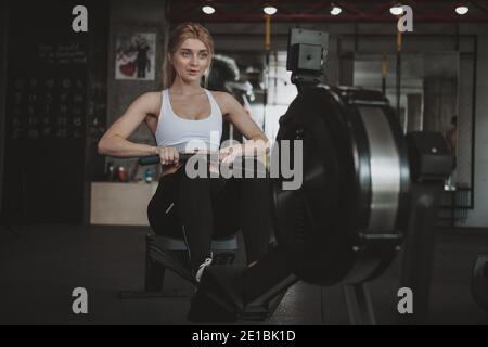 Attraente donna di fitness che guarda concentrata, esercitandosi sulla macchina di canottaggio alla palestra, spazio di copia. Sana sportivo che lavora allo studio sportivo, u Foto Stock