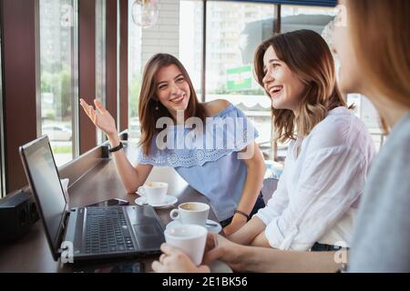 Felici e belle giovani donne che ridono, gustando il caffè del mattino insieme al caffè. Adorabili amici che chiacchierano, mentre usano il computer portatile al caffè Foto Stock