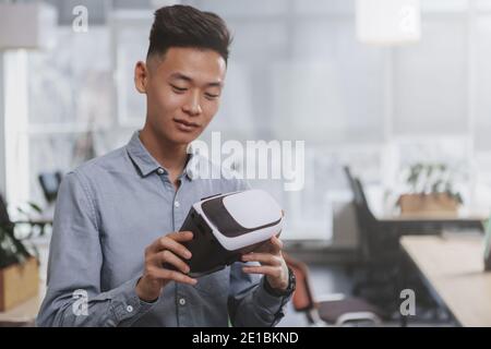 Bell'imprenditore asiatico che guarda gli occhiali 3d vr che sta tenendo. Giovane uomo d'affari che esamina le cuffie per la realtà virtuale, spazio per la copia. Sviluppo, Foto Stock