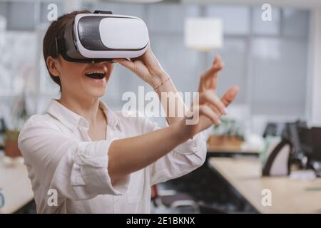 Giovane donna felice che usa gli occhiali di realtà virtuale sul lavoro, toccando qualcosa nell'aria, spazio di copia. Una donna d'affari entusiasta di utilizzare il visore 3d vr Foto Stock
