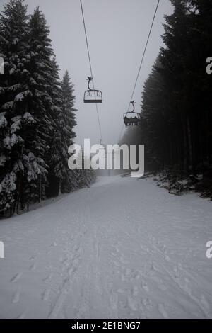 Quasi vuoto Ski Lift su un paesaggio invernale innevato, con montagne innevate e grandi pini e abeti, nella nebbia. Foto Stock