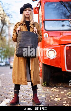 Bionda ragazza in trincea e beretto sulla strada della città con fondo autobus rosso. Giovane femmina che tiene la borsa nera. Concetto di moda o di viaggio a Londra Foto Stock