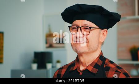Uomo anziano che indossa lo chef bonete sorridendo in cucina casalinga. Primo piano ritratto di felice vecchio panettiere pensionato con bicchieri e grembiule guardando la macchina fotografica pronta a cucinare pasticceria fatta in casa con farina e uova. Foto Stock