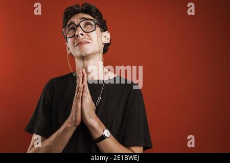 Giovane uomo pensieroso hipster che tiene le mani aggrappato e guardando in su mentre isolato su sfondo rosso Foto Stock