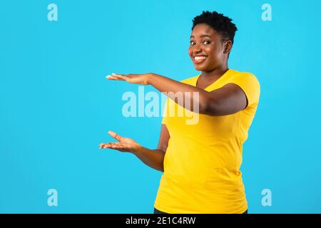 Allegro sovrappeso Africano Donna tenendo invisibile oggetto su sfondo Blu Foto Stock