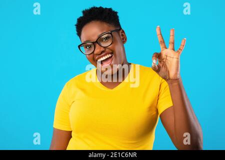 Cheerful African Plus-Size Donna gesturing okay in piedi su sfondo blu Foto Stock