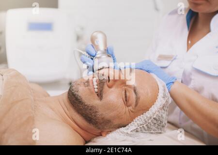 Rilassato bell'uomo maturo sorridente, ottenendo il trattamento di cavitazione ecografica da cosmetologo professionale. Estetista che usa hardware cosmetology equi Foto Stock