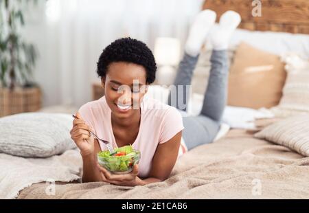 Bella signora nera mangiare insalata di verdure mentre si sdraia su comodo letto a casa, spazio copia Foto Stock