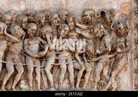 Demoni con gambe pelose e corna (a destra) trascinano i dannati nell'Inferno nel bassorilievo in marmo del giudizio universale del 1300 sul fronte occidentale del Duomo di Orvieto, Umbria, Italia. Questa scena scolpita è attribuita a Lorenzo Maitani (c. 1275 - 1330), che progettò la facciata tardo gotica della cattedrale. Si ritiene che Maitani abbia avuto un contributo diretto come scultore su due pannelli che coprono due dei quattro pilastri inferiori della facciata: Un pannello Maitani raffigura scene del giudizio universale, compresa la scena in questa immagine. Foto Stock