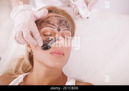 Vista dall'alto primo piano di una bella donna matura che ottiene nanogel nero applicato sul suo viso, preparandosi per il trattamento di buccia laser al carbonio Foto Stock