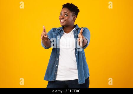 Donna Africana sorridente Plus-Size che punta le dita verso la fotocamera, sfondo giallo Foto Stock
